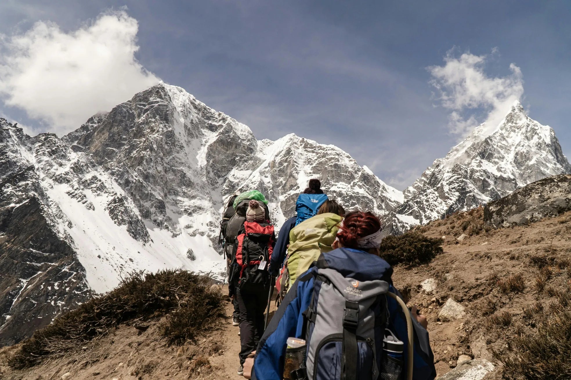 Groupe de randonneurs en montagne avec sacs à dos, progressant sur un sentier face à des sommets enneigés sous un ciel dégagé.
